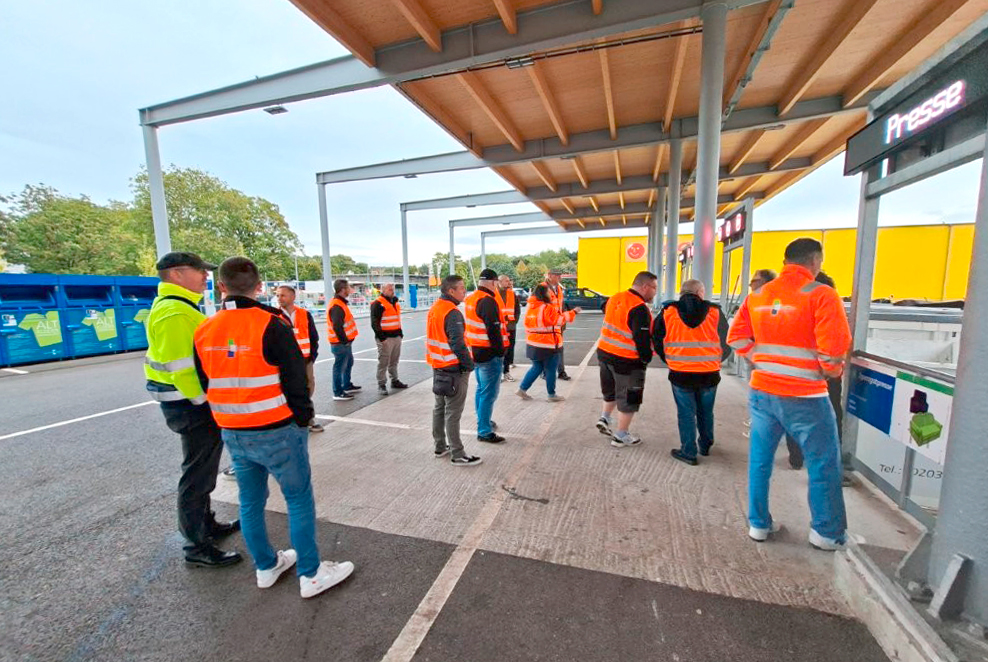 Gruppe von Personen mit orangen Warnwesten steht unter einem überdachten Bereich auf einem Recyclinghof, im Hintergrund gelbe Gebäude und blaue Container.