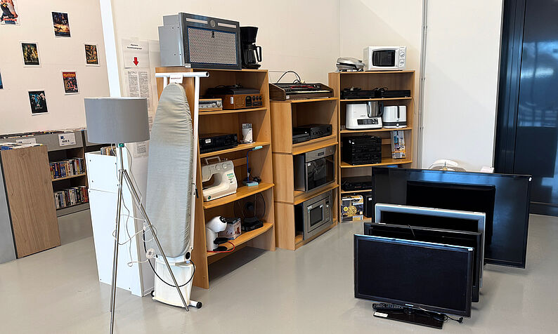 Mehrere Elektrogeräte wie Fernseher, Mikrowellen, Kaffeemaschine, Bügeleisen und ein Bügelbrett stehen in einem Raum auf dem Boden und in Regalen