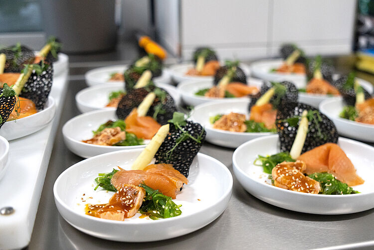 Mehrere Teller mit Lachs, Algensalat, Sesam und schwarzem Sesamcracker auf einer Küchenarbeitsplatte