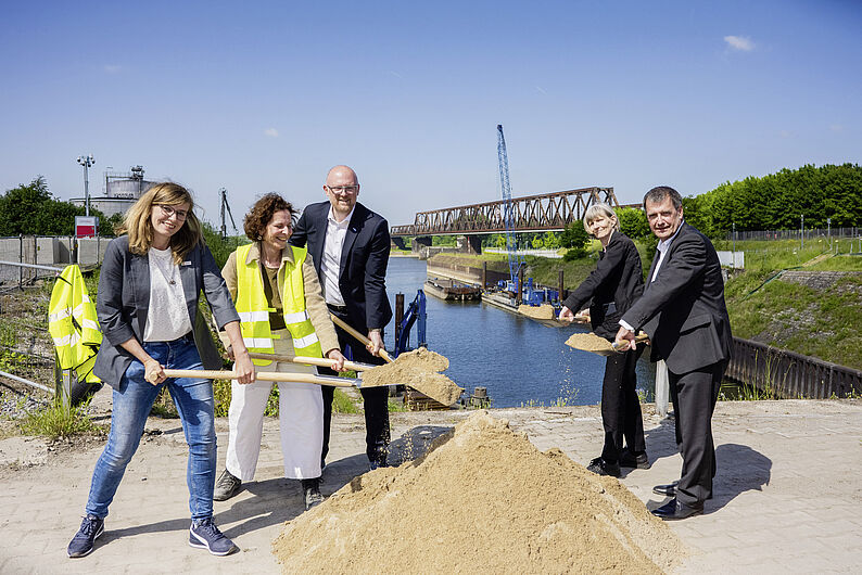 Fünf Personen mit Spaten heben Erde bei Spatenstich an einem Hafen mit Brücke im Hintergrund