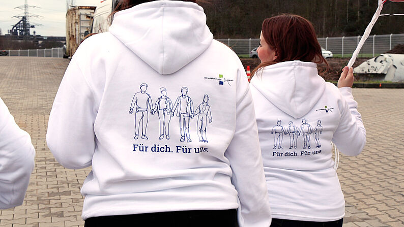 Zwei Personen tragen weiße Kapuzenpullover mit einer Illustration von vier Personen und dem Text 'Für dich. Für uns.' auf dem Rücken.