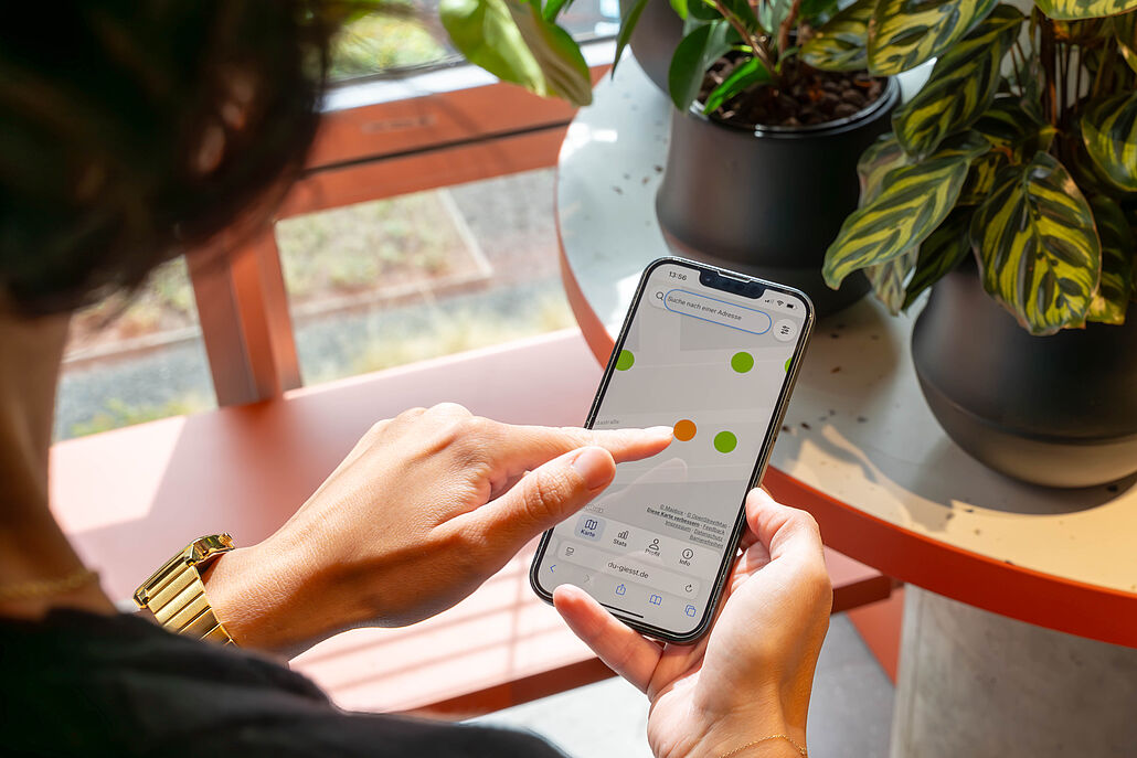 Person hält Smartphone mit einer App, die farbige Punkte anzeigt, im Hintergrund Pflanzen auf einem runden Tisch
