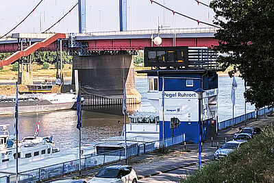 Gebäude mit der Aufschrift 'Pegel Ruhrort' am Flussufer neben einer Brücke und mehreren Booten