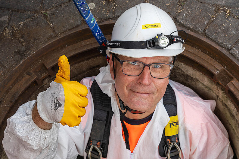 Mann in Schutzkleidung und Helm mit Stirnlampe steigt aus einem großen Gully und zeigt Daumen hoch