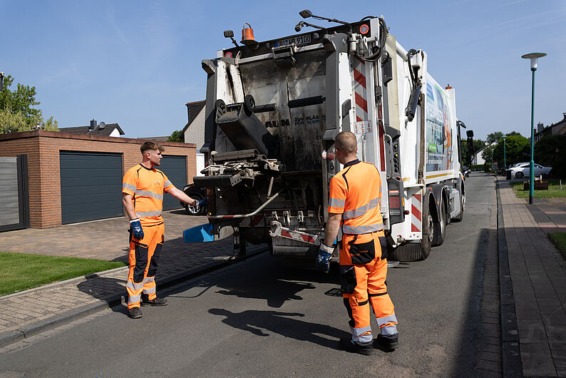 Zwei Männer der Müllabfuhr in orangefarbenen Arbeitskleidung bedienen eine Müllabfuhr auf einer Wohnstraße