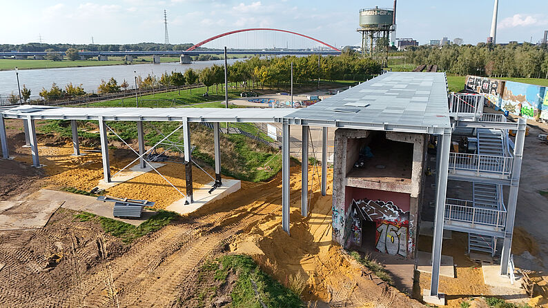 Luftaufnahme einer Baustelle mit Stahlgerüst, Betonmauer mit Graffiti und roter Brücke im Hintergrund über einem Fluss.