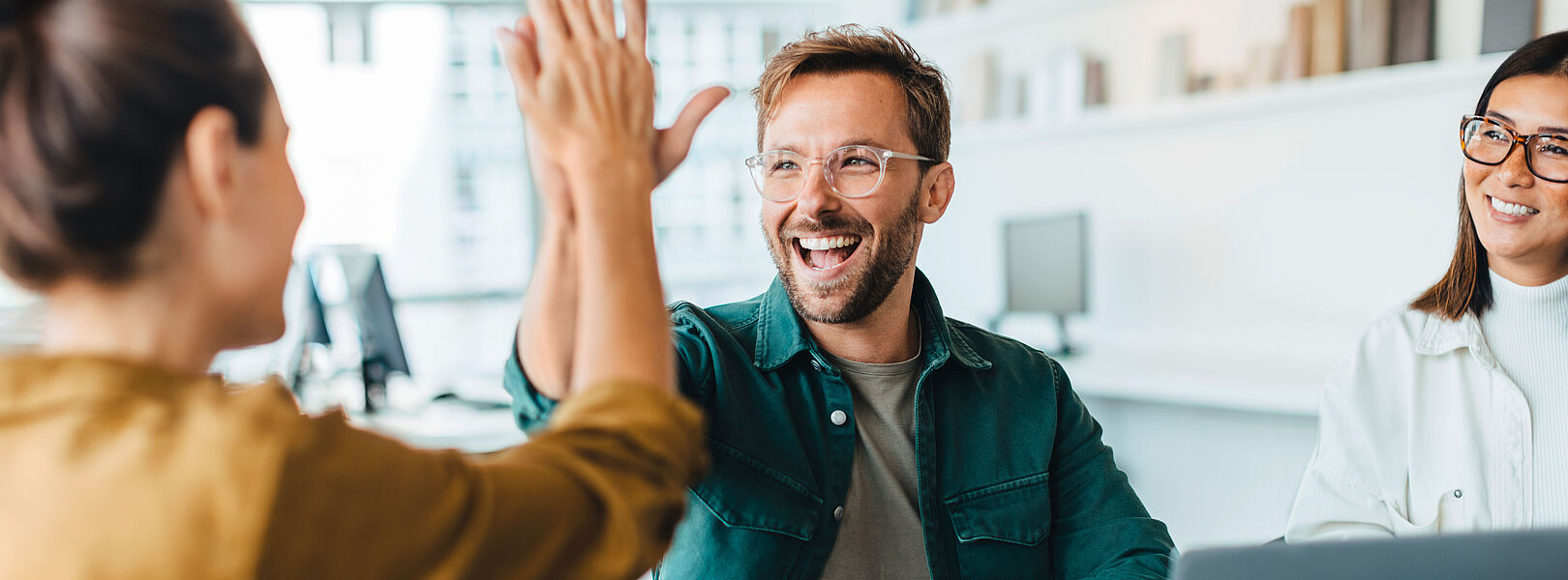 Zwei Personen geben sich im Büro einen High-Five, eine dritte Person sitzt mit Laptop am Tisch