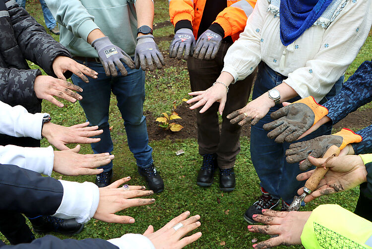 Mehrere Personen stehen auf einer Wiese im Kreis und strecken ihre mit Erde beschmutzten Hände und Arbeitshandschuhen nach innen