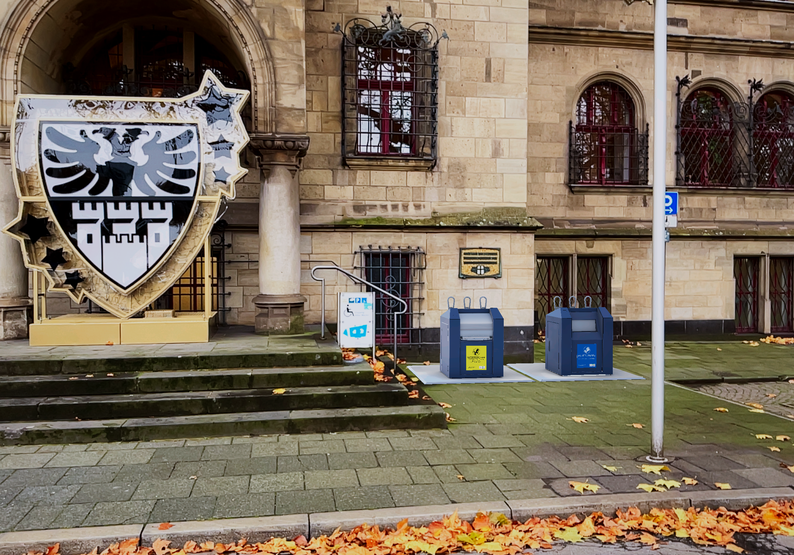 Großes Wappen mit schwarzem Adler und drei Türmen an einer steinernen Fassade neben zwei künstlich hinzugefügten blauen Müllcontainern und einem Schild mit Text.