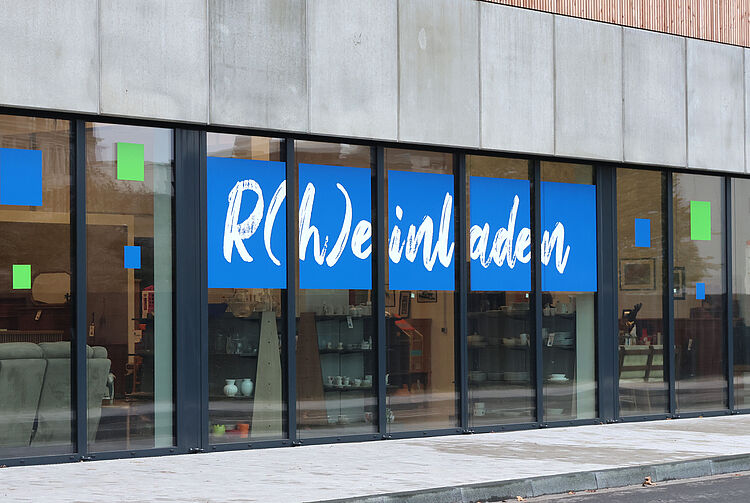 Fassade eines Ladengeschäfts mit großen Glasfenstern und Schriftzug 'R(h)einladen' in weiß auf blauem Hintergrund