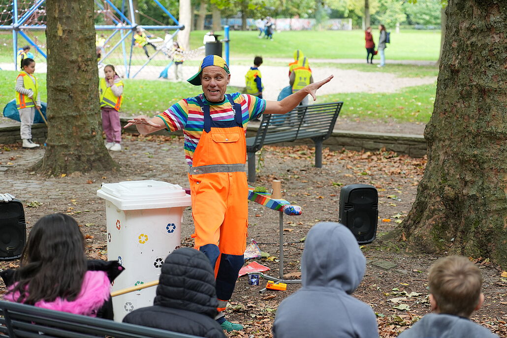 Person in orangefarbener Latzhose und gestreiftem T-Shirt steht mit ausgebreiteten Armen vor einer Gruppe sitzender Kinder im Park.