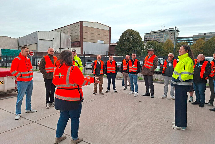Gruppe von Menschen in orangen und gelben Warnwesten steht im Halbkreis auf einem Betonplatz vor Industriegebäuden.