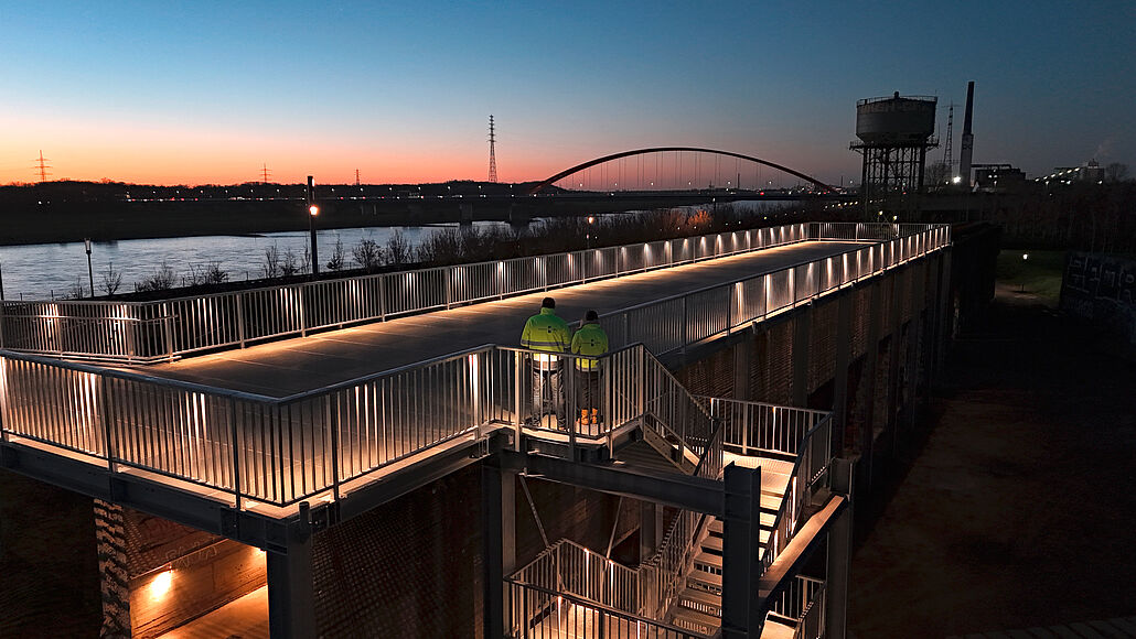 Beleuchteter Skywalk mit zwei Personen in Warnwesten bei Dämmerung. Im Hintergrundüber der Rhein mit einer Brücke 