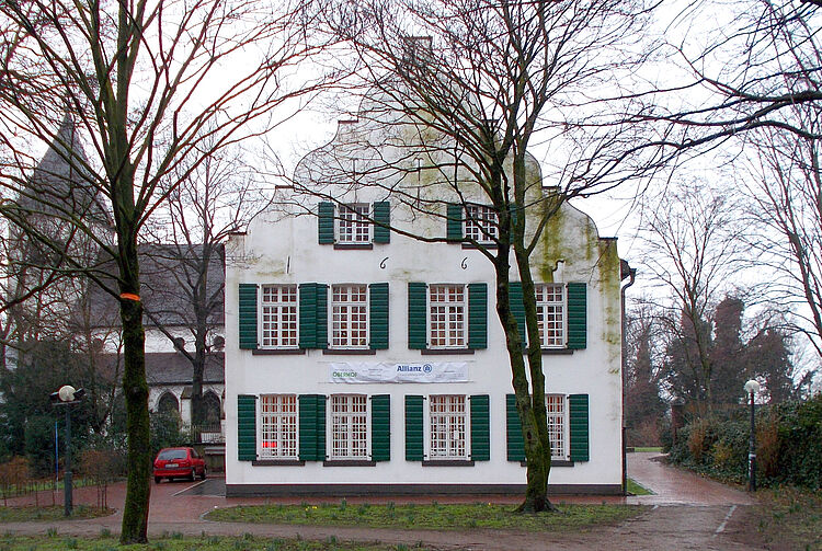 Weißes historisches Gebäude mit grünen Fensterläden und Giebel, umgeben von kahlen Bäumen und einem Weg im Vordergrund