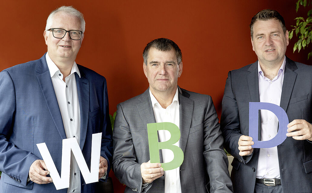 Drei Männer in Anzügen vor roter Wand, jeder hält einen Buchstaben: W, B und D. Die Männder sind Uwe Linsen, Thomas Patermann und Sebastian Beck