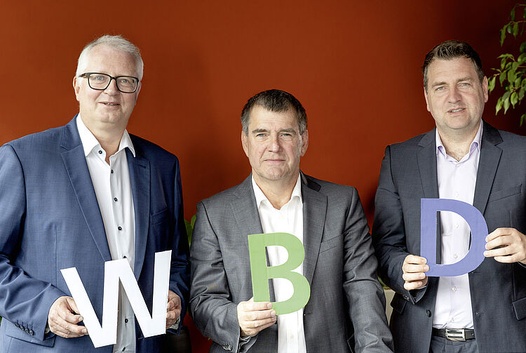Drei Männer in Anzügen vor roter Wand, jeder hält einen Buchstaben: W, B und D. Die Männder sind Uwe Linsen, Thomas Patermann und Sebastian Beck