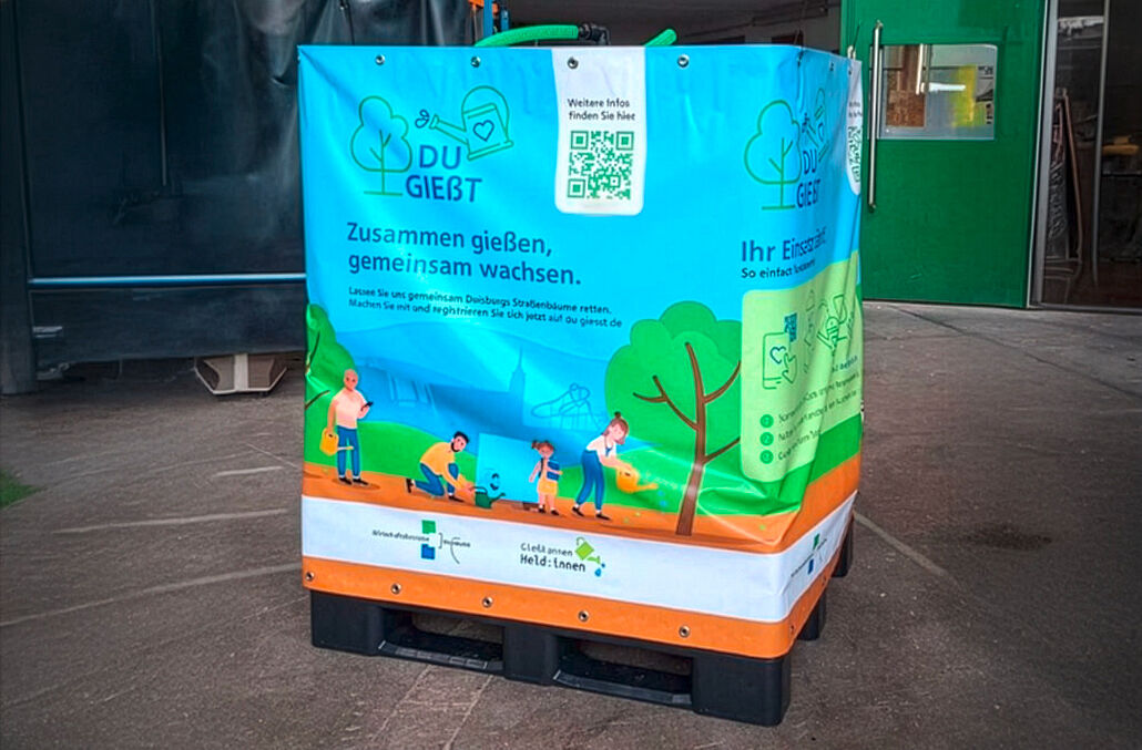 Großer blauer Wasserkanister mit Aufdruck und QR-Code auf einer schwarzen Palette in einer Halle. Headline des Aufdrucks ist "Zusammen gießen, gemeinsam wachsen."