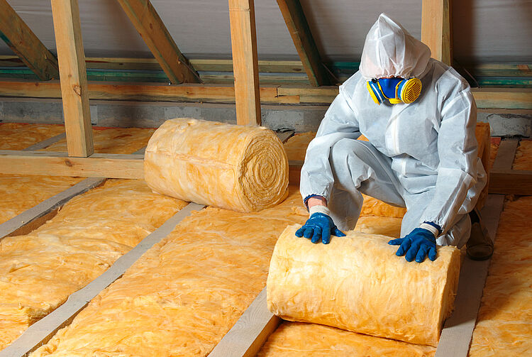 Person in Schutzanzug, Handschuhen und Atemschutzmaske verlegt Rollen aus gelber Dämmwolle zwischen Holzbalken in einem Dachboden