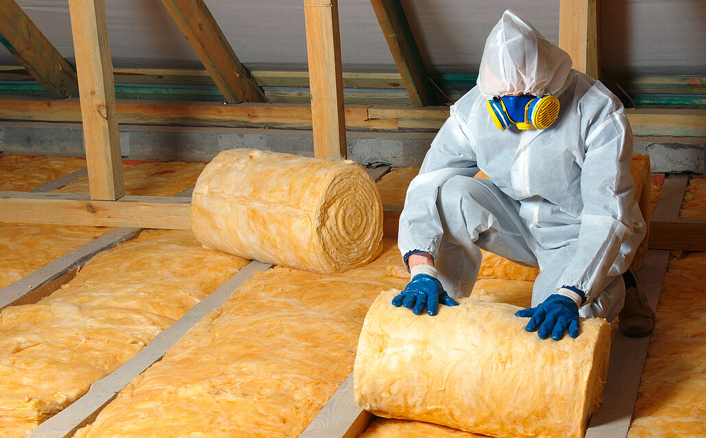 Person in Schutzanzug, Handschuhen und Atemschutzmaske verlegt Rollen aus gelber Dämmwolle zwischen Holzbalken in einem Dachboden