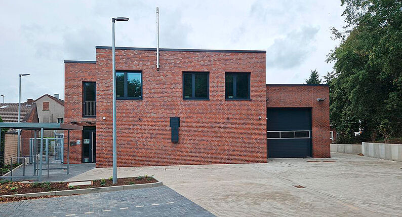 Rückseite eines zweistöckigen, roten Backsteingebäudes mit mehreren Fenstern, einer großen schwarzen Garagentür und einer Straßenlaterne davor.