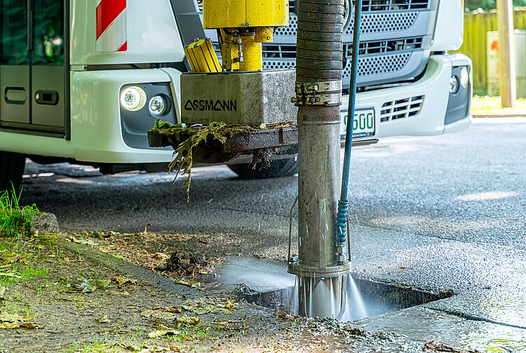 Nahaufnahme einer Maschine, die mit einem Wasserstrahl einen Gully reinigt, dahinter ein weißes Fahrzeug.