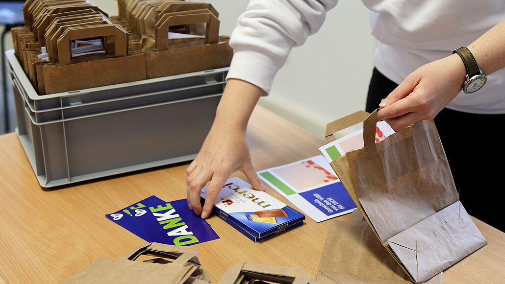 Person packt Schokoladentafel und Dankeskarte in eine braune Papiertüte auf einem Holztisch