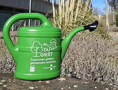 Grüne Gießkanne mit Aufdruck eines Baums, einer Gießkanne und dem Text 'DU GIEßT Zusammen gießen, gemeinsam wachsen.'