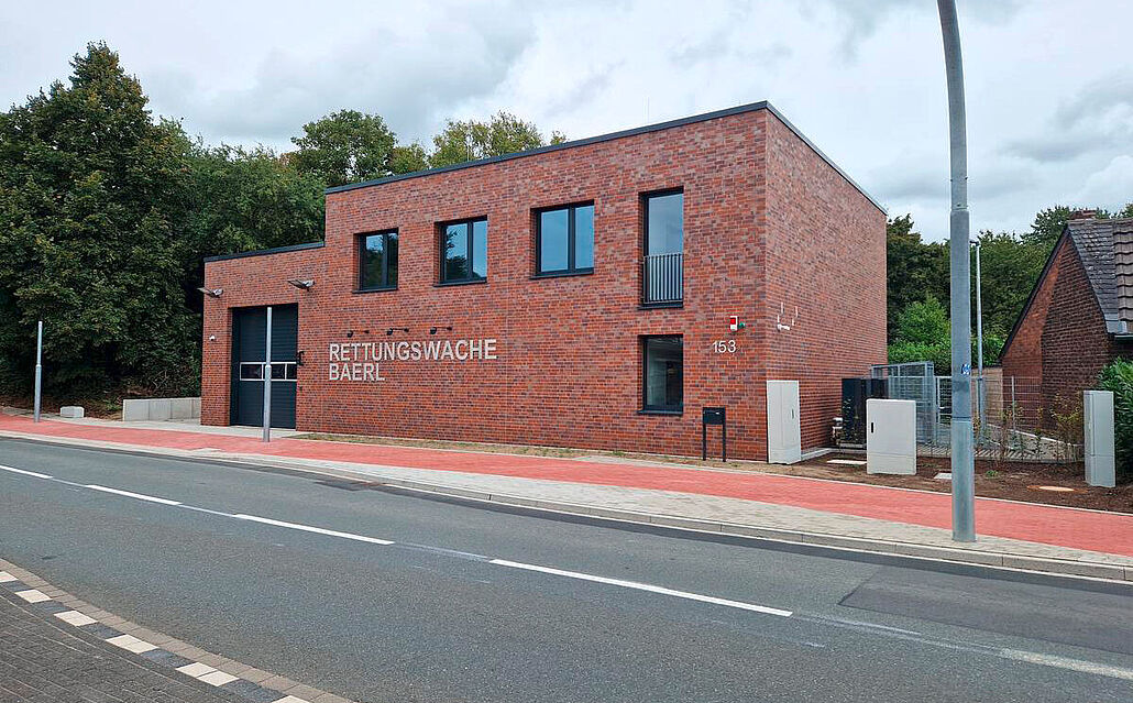 Zweistöckiges, rotes Backsteingebäude mit der Aufschrift 'RETTUNGSWACHE BAERL' und mehreren Fenstern an einer Straße.