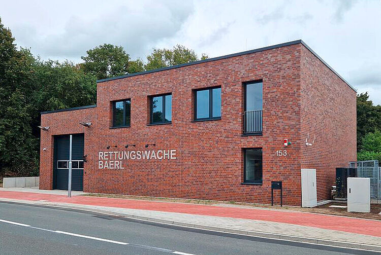 Zweistöckiges, rotes Backsteingebäude mit der Aufschrift 'RETTUNGSWACHE BAERL' und mehreren Fenstern an einer Straße.