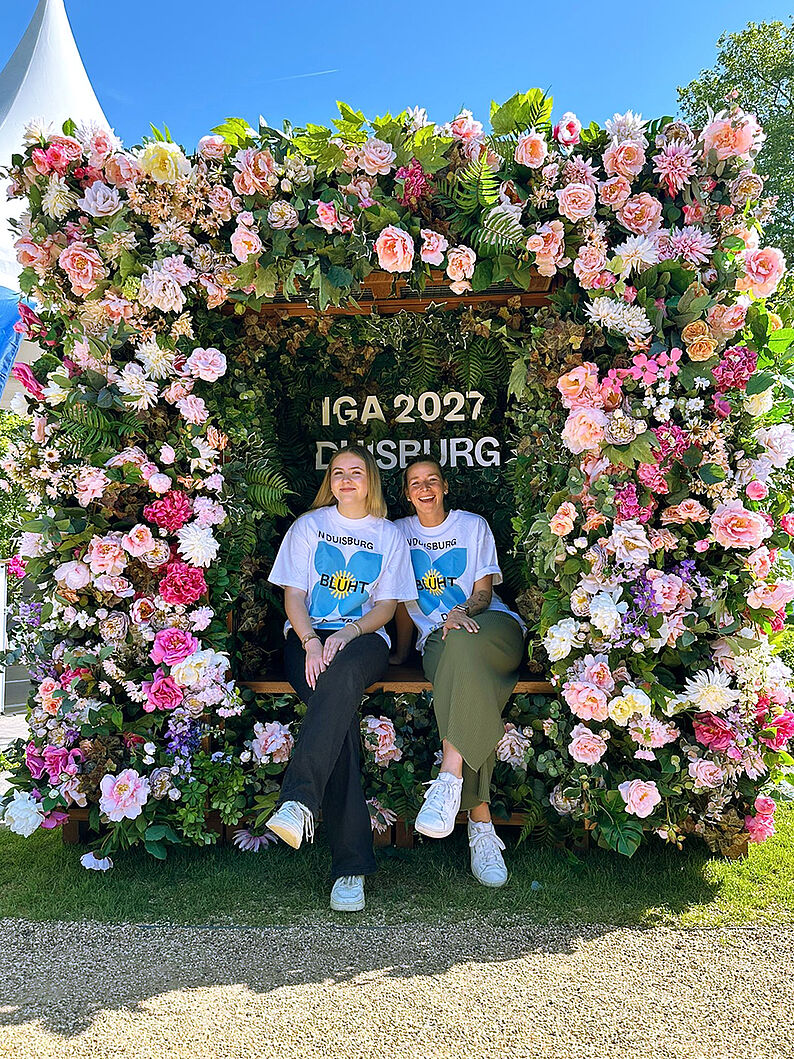 Zwei Frauen sitzen nebeneinander auf einer Bank in einem mit vielen Blumen und grünen Blättern geschmückten Selfie-Spot mit der Aufschrift 'IGA 2027 DUISBURG'