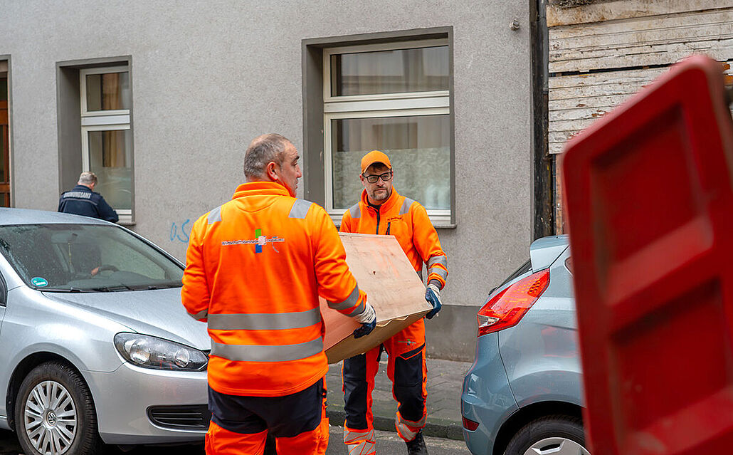 Zwei Personen in orangefarbener Arbeitskleidung tragen eine Kommode aus Holz auf einer Straße zwischen geparkten Autos