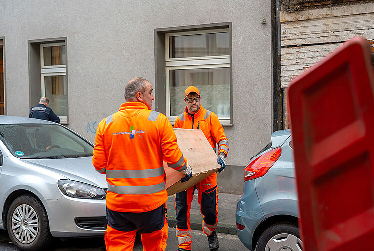 Zwei Personen in orangefarbener Arbeitskleidung tragen eine Kommode aus Holz auf einer Straße zwischen geparkten Autos