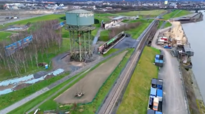 Luftaufnahme eines Industriegebiets mit einem Wasserturm, Bahngleisen, Containern und einem Flussufer