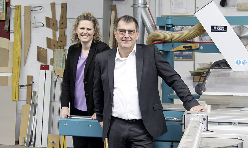 Uwe Linsen und Stephanie Schmiemann-Altenhoff, Geschäftsführerin GfB, stehen in Geschäftskleidung in einer Werkstatt neben einer großen Maschine mit Metallrohren und Werkzeugen an der Wand