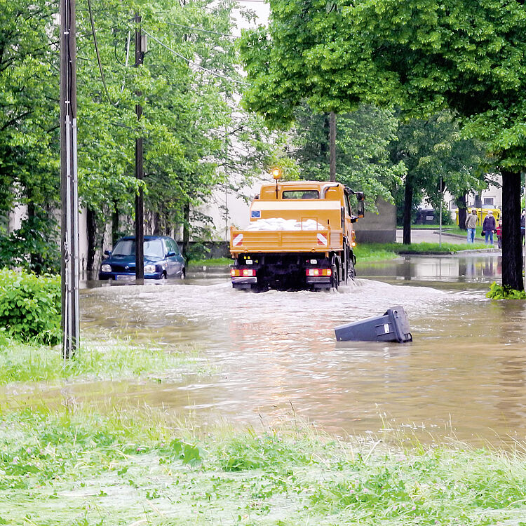 Straße mit Hochwasser überflutet, Warnschild 'Hochwasser' im Vordergrund, oranger Lastwagen fährt durch das Wasser