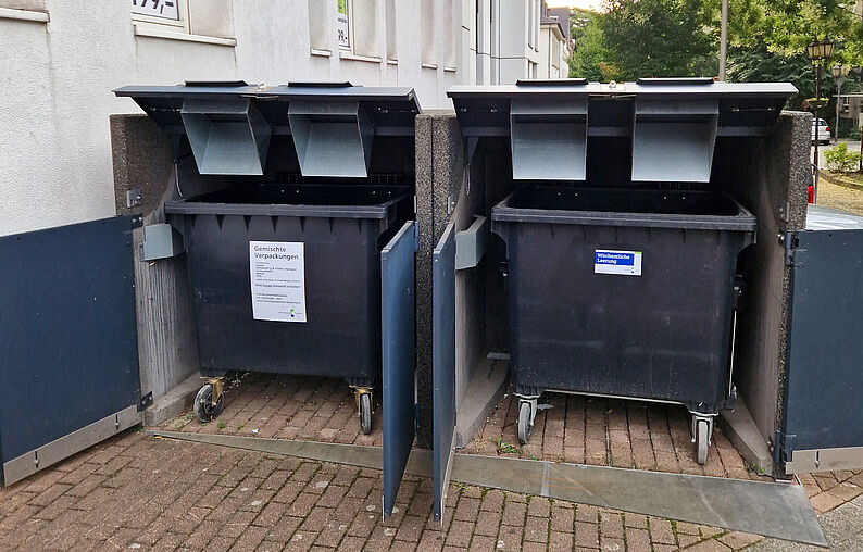Zwei große schwarze Müllcontainer mit geöffneten Deckeln in einer grauen Umhausung auf gepflastertem Boden.