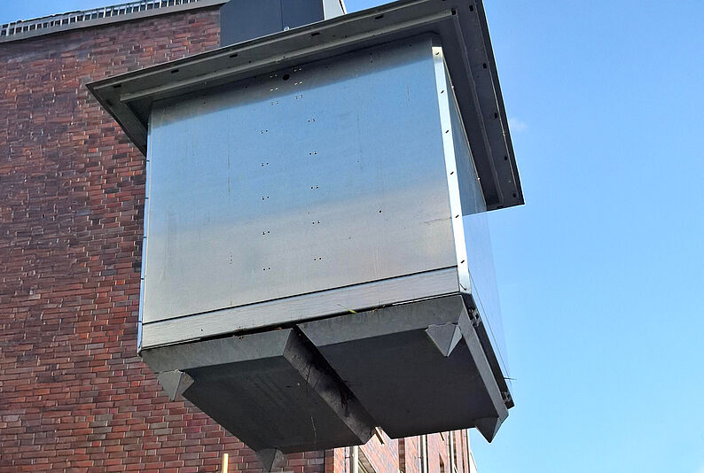 Unteransicht eines großen, rechteckigen Unterflurbehälters aus Metall vor einer roten Backsteinwand unter blauem Himmel.