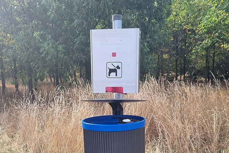 Mülleimer mit blauem Rand und Schild mit Hundesymbol zur Hundekotentsorgung in einer Wiese mit Bäumen