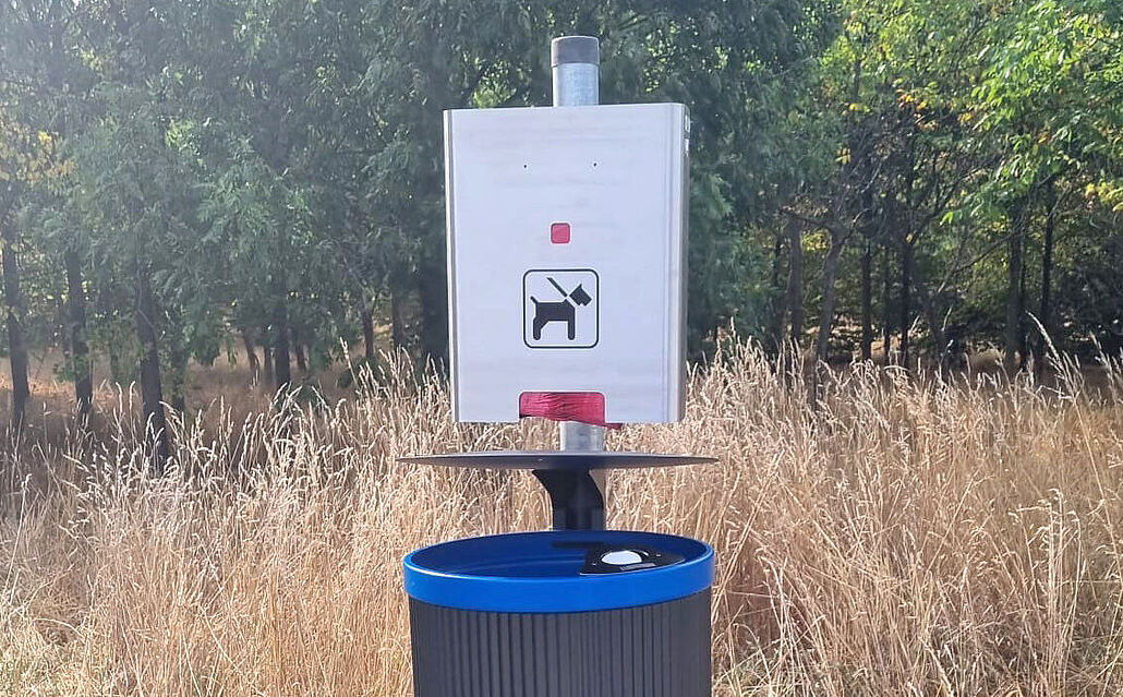 Mülleimer mit blauem Rand und Schild mit Hundesymbol zur Hundekotentsorgung in einer Wiese mit Bäumen