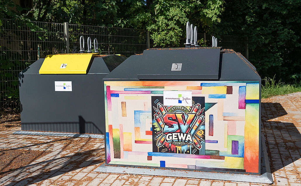 Zwei Abfall-Container auf gepflastertem Boden, einer davon mit farbiger Bemalung und Logo der Gesamtschule Walsum