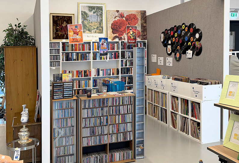 Regalwand mit zahlreichen CDs, Schallplatten und Büchern in einem Raum mit grauen Trennwänden, an der Wand eine Schallplattendekoration und gerahmte Bilder