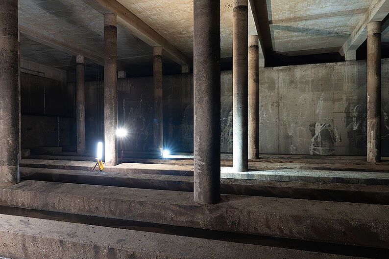 Betonraum eines Regenrückhaltebeckens mit mehreren runden Stützsäulen und zwei auf dem Boden stehenden Baustrahlern