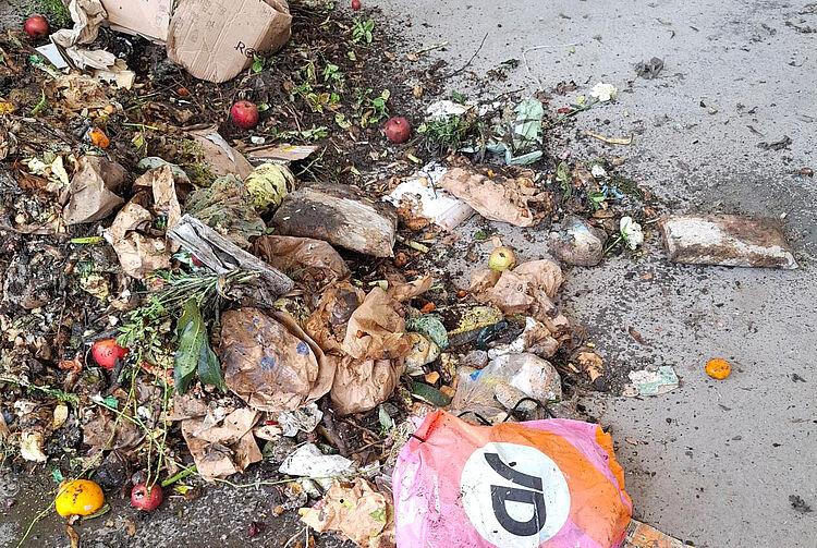 Müllhaufen mit verwelktem Gemüse, Papier, Plastik und einem pinken Beutel mit schwarzem und weißem JD-Logo auf grauem Boden.