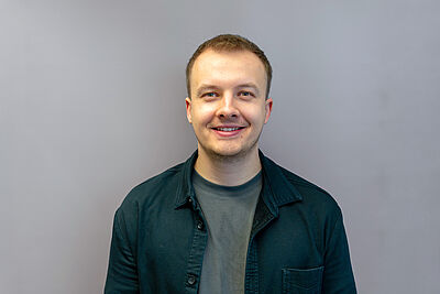 Mann mit kurzem Haar trägt ein dunkles Hemd über einem T-Shirt vor einfarbigem Hintergrund