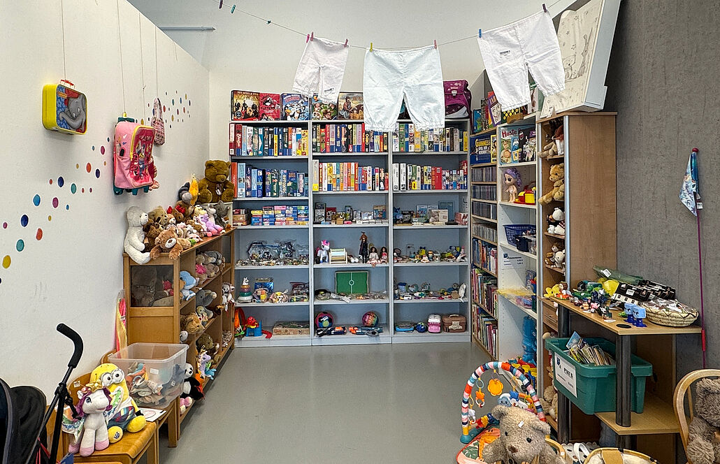 Raum aus Trennwänden mit Regalen voller Spielzeug, Kuscheltiere auf einem kleinen Tisch und Wäscheleine mit weißen Kleidungsstücken