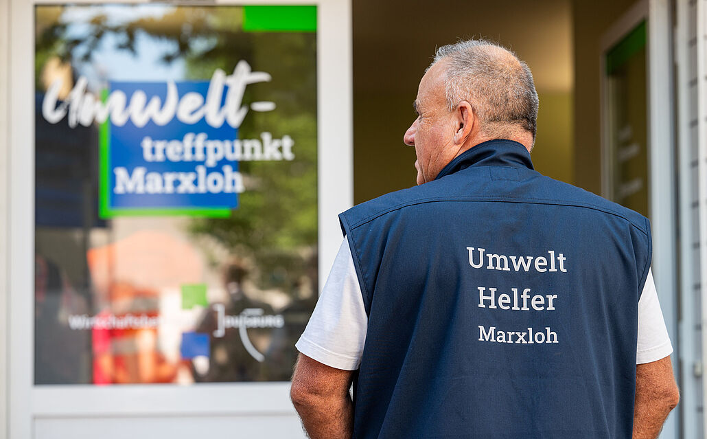 Mann mit blauem Weste mit Aufschrift 'Umwelt Helfer Marxloh' steht vor Tür mit Schild 'Umwelt-treffpunkt Marxloh'.
