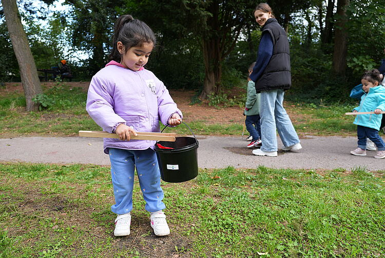 Kind in lila Jacke und blauer Jeans hält einen schwarzen Eimer und eine Müllzange auf einer Wiese, im Hintergrund weitere Personen auf einem Weg.