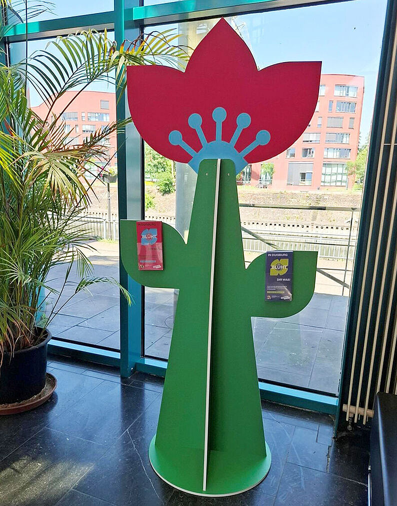 Grüner Blumenaufsteller mit roter Blüte und zwei angebrachten Broschüren