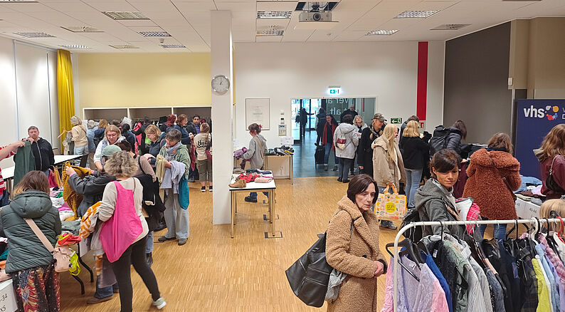 Mehrere Personen in einem Raum mit Garderobenständern und Tischen, auf denen Kleidung liegt, bei einer Altkleidersammlung.