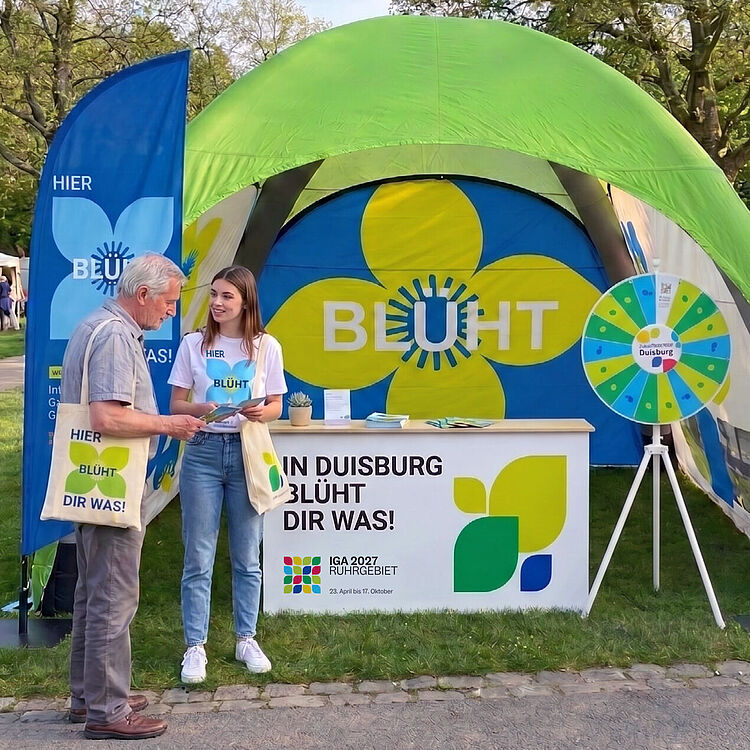 Zelt mit grünem Dach und Werbestand mit gelber Blume und Schriftzug "BLÜHT" sowie zwei Personen, die sich unterhalten, umgeben von blauen und roten Fahnen mit demselben Schriftzug